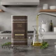 Premium Koroneiki variety olive oil being poured into a glass carafe on a marble countertop next to fresh rosemary and peppercorns.