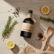 Top-down view of Navarino Icons olive oil bottle surrounded by fresh lemons, rosemary, peppercorns, and wooden spoons on a light wood surface.